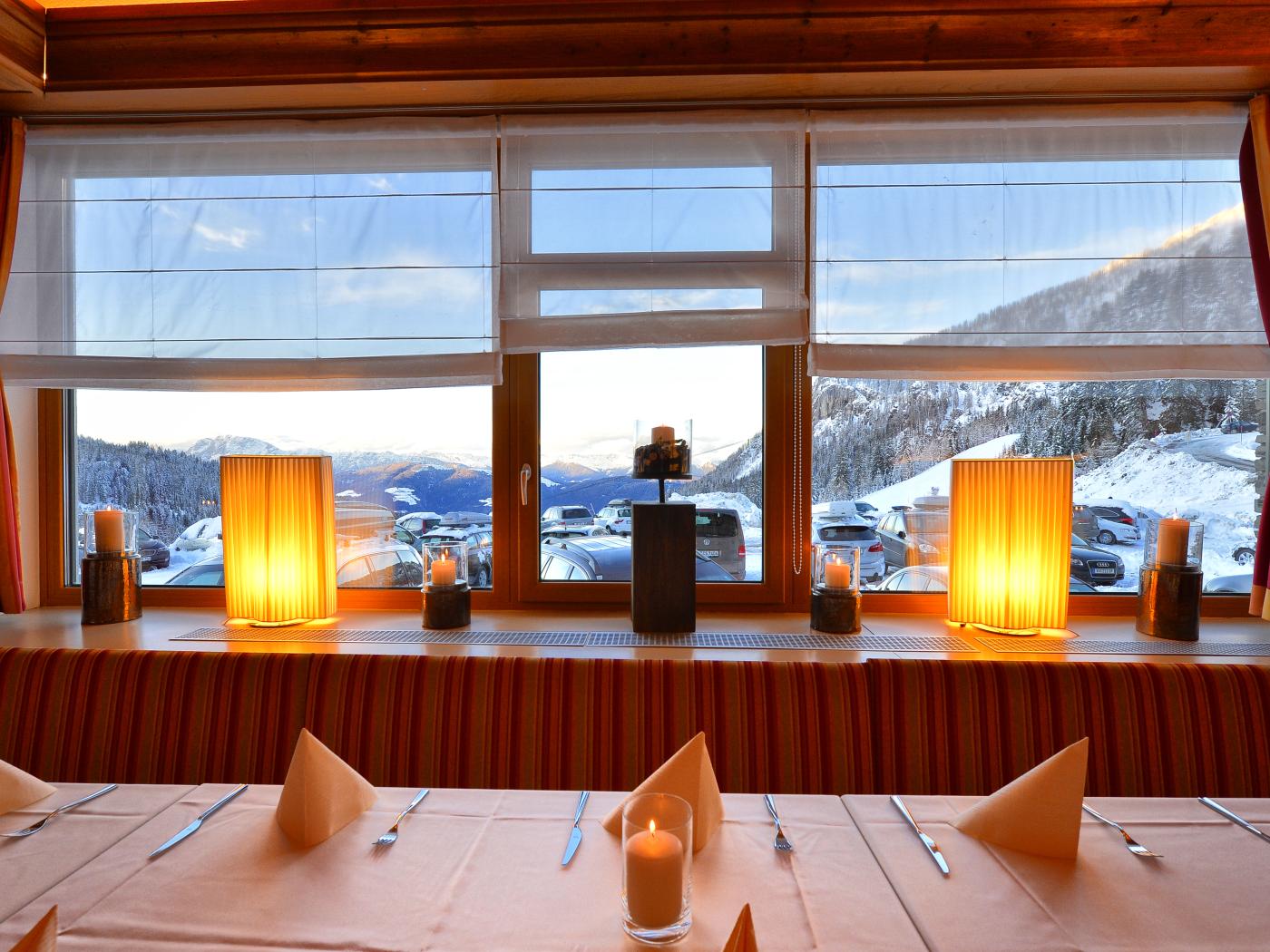 Ein stilvoll gedeckter Tisch im Almhotel Kärnten mit Blick auf verschneite Berge und untergehende Sonne. Warme Lampen sorgen für eine einladende Atmosphäre, während das Panoramafenster eine herrliche Aussicht auf die Winterlandschaft der Kärntner Alpen bietet. Gemütliches Restaurant mit Bergblick im Almhotel Kärnten am Nassfeld