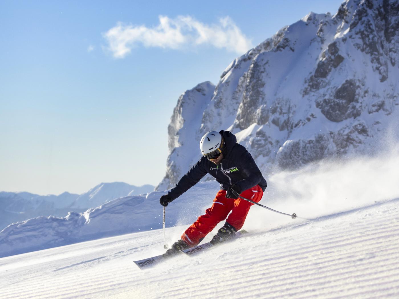 Ein Skifahrer gleitet schwungvoll über eine frisch präparierte Piste vor beeindruckender Alpenkulisse. Die Sonnenstrahlen lassen den Schnee glitzern, während der tiefblaue Himmel und majestätische Felswände für eine traumhafte Winterstimmung sorgen. Ideale Bedingungen für Wintersport in Kärnten. Skifahren auf perfekt präparierter Piste am Nassfeld in der Nähe des Almhotel Kärnten