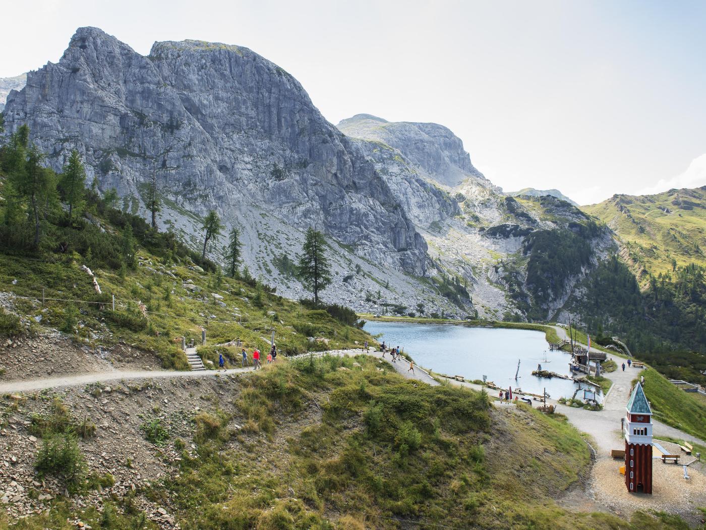 Ein idyllischer Bergsee im Naturparadies Kärnten, umgeben von beeindruckenden Felswänden und grüner Vegetation. Ein Wanderweg führt entlang des klaren Sees, an dessen Ufer ein kleiner Steg und ein charakteristischer roter Turm zu sehen sind. Entspannung und Naturerlebnis pur. Aqua-Trail Familien-Erlebnisweg am Nassfeld, in der Nähe des Almhotel Kärnten