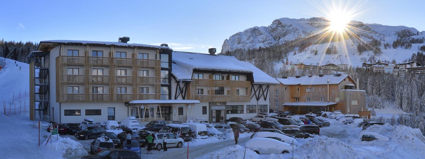 Das Almhotel Kärnten präsentiert sich in winterlicher Pracht am Fuß verschneiter Berge. Die Sonne geht hinter einer Felsformation auf und beleuchtet das moderne Hotelgebäude. Skifahrer und Wintersportler genießen die direkte Lage in der verschneiten Natur der Karnischen Alpen. Ski-In und Ski-Out im Almhotel Kärnten am Nassfeld direkt an der Skipiste