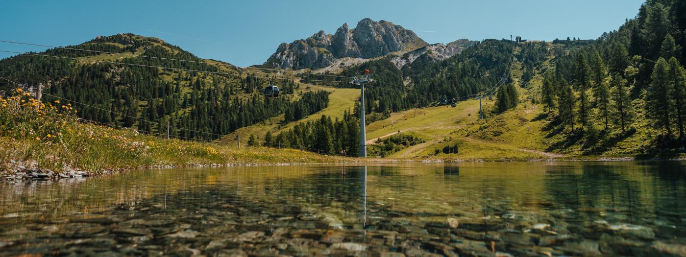 Der kristallklare Watschiger Almsee am Nassfeld mit Blick auf grüne Almwiesen, Wälder und die markanten Gipfel des Gartnerkofels. Die ruhige Wasseroberfläche und das klare Sommerlicht vermitteln alpine Erholung in Kärnten. Watschiger Almsee am Gartnerkofel mit Gartnerkofelbahn