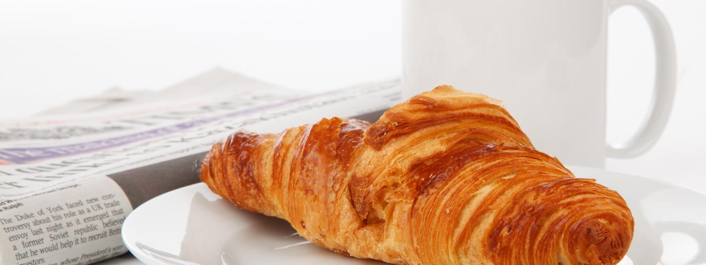 Knuspriges Croissant auf einem weißen Teller, daneben eine Tasse Kaffee und eine Tageszeitung. Ein perfekter Start in den Tag im Almhotel Kärnten, ideal für Genießer eines entspannten Frühstücks in gemütlicher Atmosphäre. Frisches Croissant mit Kaffee im Almhotel Kärnten am Nassfeld