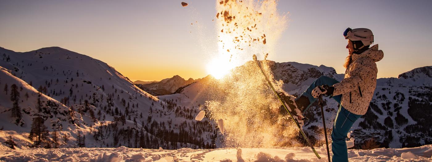 Eine Skifahrerin genießt den Sonnenuntergang in den verschneiten Karnischen Alpen. Sie wirbelt frischen Schnee auf, während goldenes Licht die Landschaft erhellt. Das winterliche Panorama und die fröhliche Stimmung laden zu unvergesslichen Urlaubsmomenten ein. Ski- und Winterspaß im Almhotel Kärnten am Nassfeld