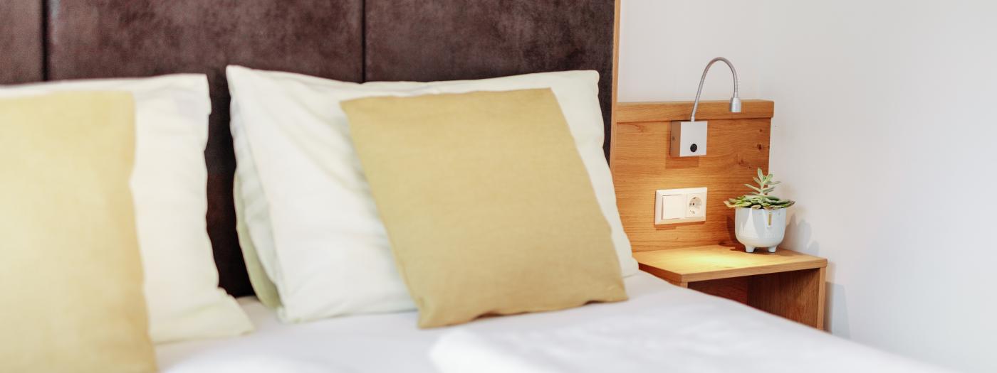 Modernes Hotelzimmer mit komfortablem Doppelbett, hellen Kissen und weicher Bettwäsche. Das Nachttischchen aus Holz ist mit einer kleinen Pflanze und einer Leselampe dekoriert. Die gemütliche Atmosphäre lädt zum Entspannen im Almhotel Kärnten ein. Hell eingerichtetes Hotelzimmer im Almhotel Kärnten am Nassfeld