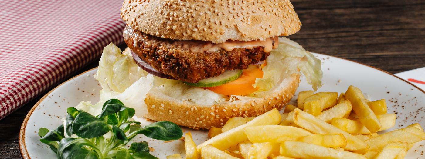 Ein leckerer, knuspriger Burger mit Sesambrötchen, knackigem Salat, Tomate und einer herzhaften Sauce, serviert mit goldgelben Pommes Frites und frischem Vogerlsalat. Das ansprechende Arrangement auf rustikalem Holztisch lädt zum Genuss ein. Knuspriger Burger mit Pommes Frites - serviert in der Garnitzenalm, in der Nähe des Almhotel Kärnten am Nassfeld