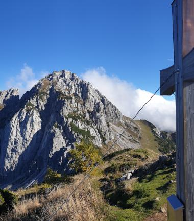 Beeindruckende Panoramaaufnahme vom Gipfel eines Kärntner Berges mit markantem Gipfelkreuz im Vordergrund. Im Hintergrund erheben sich schroffe Felshänge und das Alpenpanorama unter strahlend blauem Himmel. Die klare Sicht und das natürliche Licht schaffen eine majestätische, ruhige Atmosphäre. Panoramablick auf den Gartnerkofel-Westgrat vom Kammleiten-Gipfelkreuz am Nassfeld aus