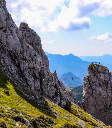 Im Vordergrund beeindruckende, steile Felsformationen, dahinter weitläufige grüne Berghänge und eine markante Felsspitze mit Besuchern auf dem Gipfel. Klare Fernsicht bis zu den blauen Alpen. Perfekte Kulisse für Wanderer und Bergliebhaber in Kärnten. Klettersteig Däumling am Gartnerkofel am Nassfeld mit imposanter Nepal-Brücke