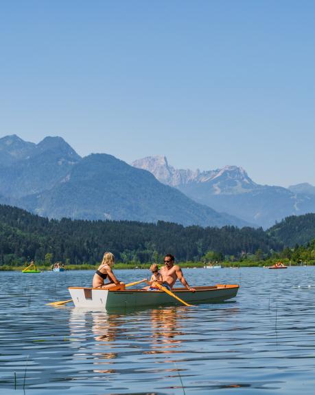 Ein Paar genießt sommerliches Wetter auf einem klaren See in Kärnten und rudert gemeinsam mitten in alpiner Naturlandschaft. Im Hintergrund erheben sich majestätische Berge und bewaldete Ufer, die das idyllische Ambiente unterstreichen. Perfekte Szene für Natur- und Aktivurlauber. Ruderboot am Pressegger See in der Region Nassfeld in Kärnten in der Nähe des Almhotel Kärnten