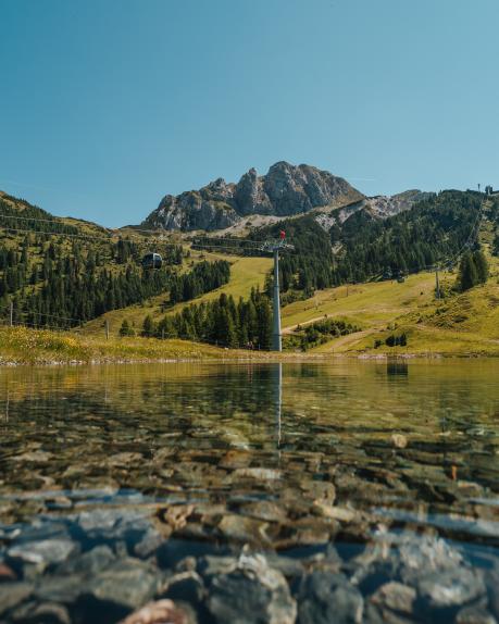 Der kristallklare Watschiger Almsee am Nassfeld mit Blick auf grüne Almwiesen, Wälder und die markanten Gipfel des Gartnerkofels. Die ruhige Wasseroberfläche und das klare Sommerlicht vermitteln alpine Erholung in Kärnten. Watschiger Almsee am Gartnerkofel mit Gartnerkofelbahn