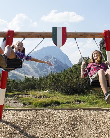 Zwei Kinder schaukeln lachend auf einer bunten Holzschaukel inmitten der Kärntner Alpen. Die Berge und der blaue Himmel im Hintergrund unterstreichen das naturnahe und fröhliche Ambiente einer unbeschwerten Auszeit in der Natur. Grenz-Schaukel am Dolce Vita Weg am Nassfeld nahe dem Almhotel Kärnten