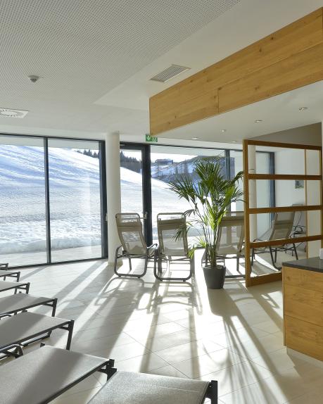 Moderner Ruhebereich mit bequemen Liegestühlen und großen Fensterfronten, die einen beeindruckenden Blick auf die verschneite Winterlandschaft bieten. Helle Holzelemente und stilvolle Pflanzen sorgen für eine entspannte, natürliche Atmosphäre im Almhotel Kärnten. Ideal zum Ausruhen nach einem Tag im Schnee. Ruheraum der Saunawelt mit Panoramablick auf die Piste im Almhotel Kärnten am Nassfeld