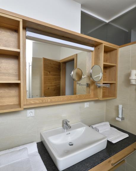Ein stilvoll eingerichtetes Hotelbad mit geräumigem Waschbecken, großem Spiegel und offenen Holzregalen. Auf dem Waschtisch liegen frische Handtücher bereit, ein Haartrockner und Kosmetikspiegel bieten zusätzlichen Komfort. Helle Fliesen schaffen eine freundliche und einladende Atmosphäre. Moderne Badezimmer in den Hotelzimmern im Almhotel Kärnten am Nassfeld