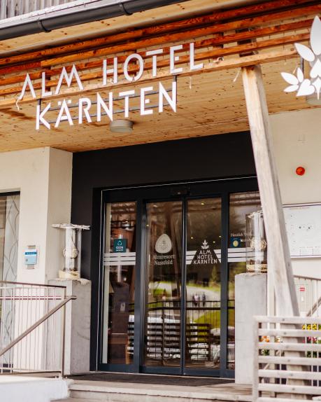 Der moderne Eingangsbereich des Almhotels Kärnten präsentiert sich mit einer stilvollen Holzfassade und klarer Beschilderung. Große Glasfronten bieten einen einladenden Blick ins Innere. Die harmonische Architektur verbindet alpinen Charme mit zeitgemäßem Design im Herzen Kärntens. Eingangsbereich des Almhotels Kärnten am Nassfeld