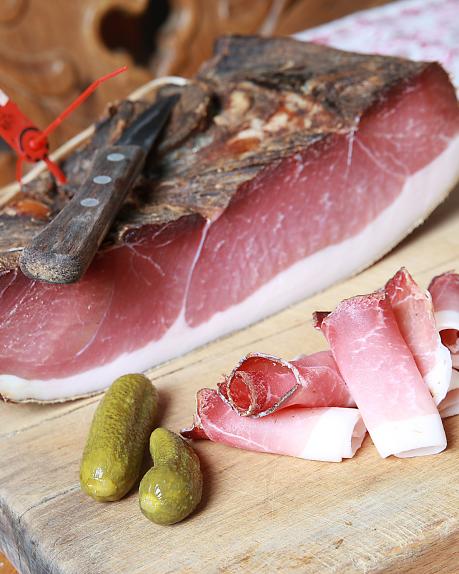 Hausgemachter Kärntner Bauernspeck, kunstvoll auf einem rustikalen Holzbrett angerichtet. Neben fein geschnittenen Speckstreifen liegen zwei knackige Essiggurkerl. Im Hintergrund sorgen traditionelle Elemente für authentisches Almhütten-Flair. Original Gailtaler Speck kann im Almhotel Kärnten am Nassfeld genossen werden
