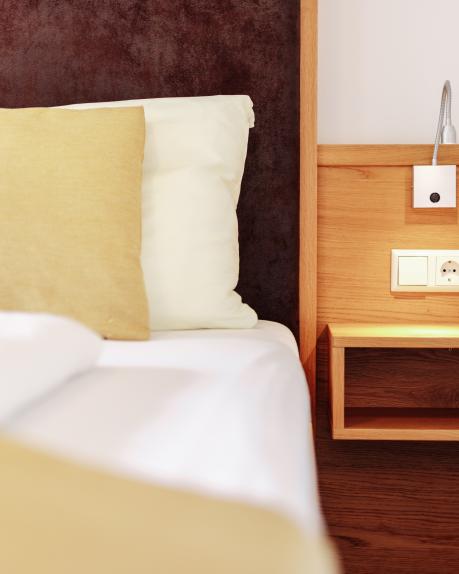 Detailaufnahme eines stilvollen Hotelzimmers mit weichem Kopfkissen, moderner Leseleuchte und einem Nachttisch aus Holz. Eine kleine grüne Pflanze sorgt für einen frischen Akzent. Perfekte Wohlfühlatmosphäre für entspannte Nächte im Almhotel Kärnten. Gemütliches Hotelzimmer mit Nachttisch im Almhotel Kärnten am Nassfeld