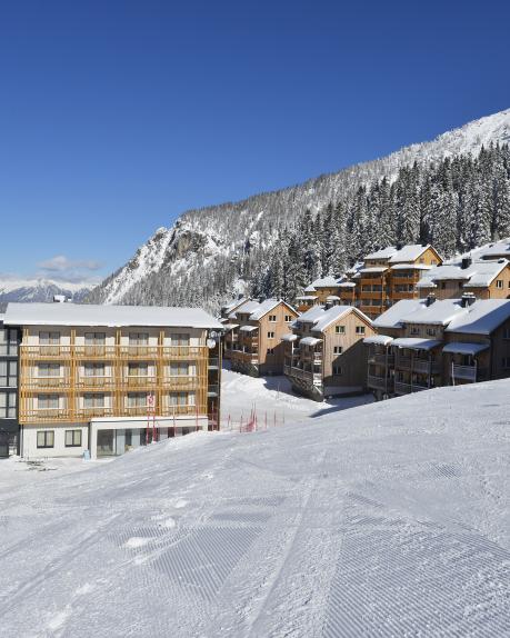 Das Almhotel Kärnten liegt eingebettet in eine verschneite Berglandschaft direkt an der Skipiste. Moderne Holzarchitektur, traditionelle Chalets und eine malerische Winterkulisse mit schneebedecktem Hang und umliegendem Nadelwald bieten perfekte Bedingungen für einen Skiurlaub in Kärnten. Das Almhotel Kärnten liegt direkt an der Piste im Skigebiet Nassfeld