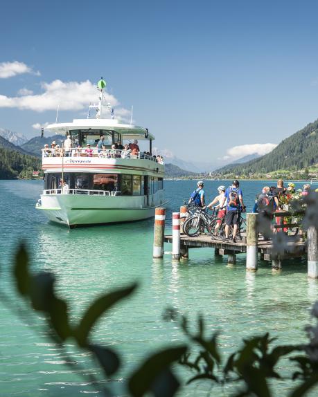 Ein Ausflugsschiff legt an einem sonnigen Tag am Steg des Weissensees an. Gäste mit Fahrrädern und Familien warten auf das Boarding, umgeben von türkisblauem Wasser und malerischen Bergen. Die idyllische Kulisse in Kärnten lädt zum Entspannen und Genießen ein. Schifffahrt auf dem Weissensee - Ausflugstipp für Gäste des Almhotel Kärnten am Nassfeld