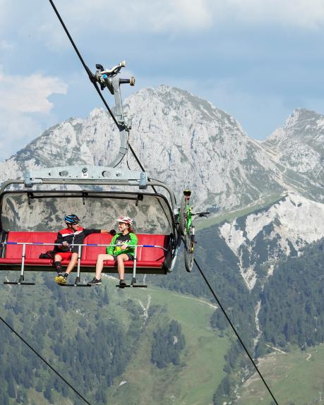 Zwei Personen fahren entspannt in einer modernen Sesselbahn mit Fahrrad-Halterung über grüne Berghänge. Im Hintergrund erhebt sich eindrucksvoll ein markanter Gipfel der Kärntner Alpen unter blauem Himmel, ideal für Mountainbiker und Naturliebhaber. Per Madritschen 6-er Sesselbahn am Nassfeld gelangen Biker auf den Berg