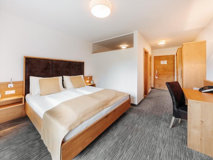 Helles und komfortables Doppelzimmer im Almhotel Kärnten mit großzügigem Bett, stilvollen Holzmöbeln und Schreibtisch. Sanfte Farben und viel Tageslicht sorgen für eine entspannte Atmosphäre – ideal für erholsame Urlaube in ruhiger Umgebung. Modernes, einladendes Doppelzimmer mit Holzmöbeln im Almhotel Kärnten am Nassfeld