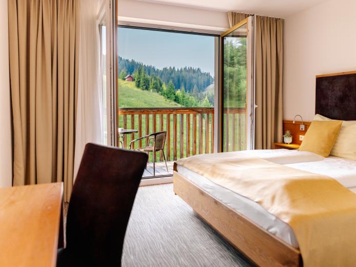Modern eingerichtetes Doppelzimmer mit großem Fenster und Balkon, das einen herrlichen Blick auf die umliegenden grünen Wiesen und Wälder bietet. Helle Farben und natürliche Materialien schaffen eine entspannte Wohlfühlatmosphäre – perfekt für einen erholsamen Aufenthalt im Almhotel Kärnten. Komfortables Doppelzimmer mit Bergblick im Almhotel Kärnten am Nassfeld