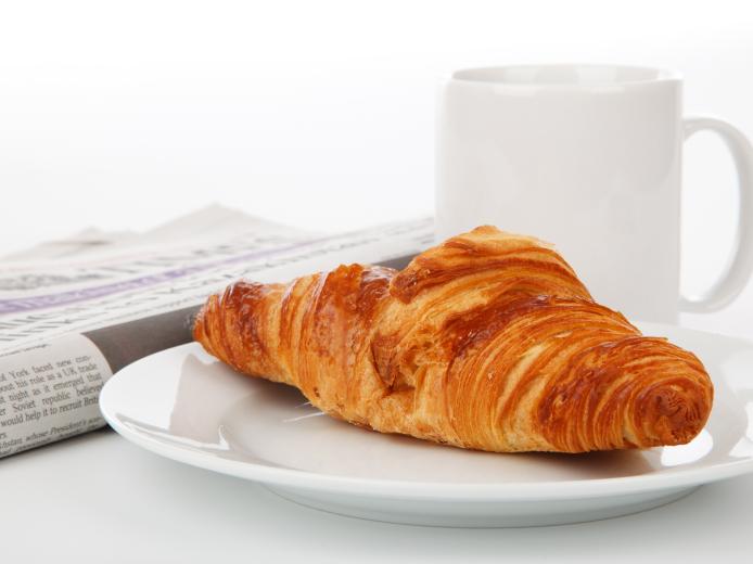 Knuspriges Croissant auf einem weißen Teller, daneben eine Tasse Kaffee und eine Tageszeitung. Ein perfekter Start in den Tag im Almhotel Kärnten, ideal für Genießer eines entspannten Frühstücks in gemütlicher Atmosphäre. Frisches Croissant mit Kaffee im Almhotel Kärnten am Nassfeld