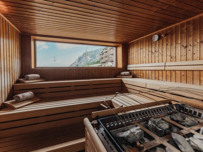 Helles, komfortables Saunazimmer mit großen Holzsitzbänken und modernem Saunaofen. Das breite Panoramafenster bietet einen beeindruckenden Blick auf die umliegenden Berge und lässt viel Tageslicht herein. Perfekt für entspannte Wellnessmomente im Almhotel Kärnten. Gemütliche Panoramasauna mit Ausblick in der Saunawelt des Almhotel Kärnten am Nassfeld