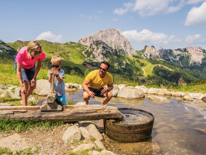 Eine Familie genießt einen sonnigen Tag an einem klaren Bergsee in Kärnten. Kinder spielen an einem kleinen Holzbrunnen mit frischem Quellwasser, während grüne Almwiesen und majestätische Gipfel die idyllische Berglandschaft umrahmen. Perfekter Ort für Naturerlebnisse und Entspannung. Der Aqua-Trail am Nassfeld in Kärnten ist bequem vom Almhotel Kärnten aus erreichbar