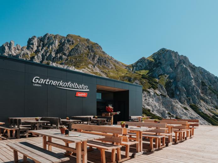 Sonnige Holzterrasse bei der Gartnerkofelbahn Bergstation am Nassfeld mit rustikalen Sitzbänken und Blick auf die markanten Felsgipfel der Kärntner Alpen. Die klare Bergluft und alpine Kulisse schaffen eine einladende Atmosphäre für eine Sommerpause. Gartnerkofelbahn Bergstation - Sonnenterrasse der Einkehrhütte Berghex im Sommer