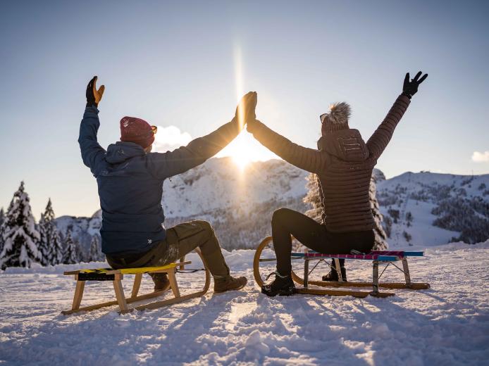 Zwei Menschen sitzen auf Schlitten im verschneiten Winterparadies und geben sich zum Sonnenuntergang ein High Five. Im Hintergrund erheben sich schneebedeckte Berge, die für eine ausgelassene, fröhliche Stimmung und perfekte Wintererlebnisse sorgen. Rodelspaß am Nassfeld in der Nähe des Almhotel Kärnten