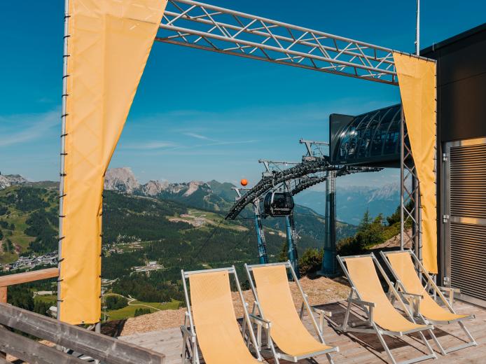 Sonnige Terrasse bei der Bergstation der Gartnerkofelbahn am Nassfeld mit Liegestühlen, Holzdeck und weitem Blick auf die Kärntner Bergwelt. Die Seilbahnanlage und der klare Himmel vermitteln alpines Sommerflair nahe dem Almhotel Kärnten. Sonnenterrasse bei der Bergstation der Gartnerkofelbahn