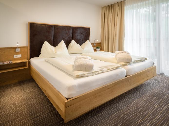 Helles und gemütliches Doppelzimmer mit komfortablem Doppelbett, modernen Holzmöbeln und direktem Zugang zum Balkon. Große Fenster sorgen für viel Tageslicht, während weiche Bettwäsche und kuschlige Handtücher für Wohlfühlatmosphäre im Almhotel Kärnten sorgen. Modernes Doppelzimmer im Almhotel Kärnten am Nassfeld