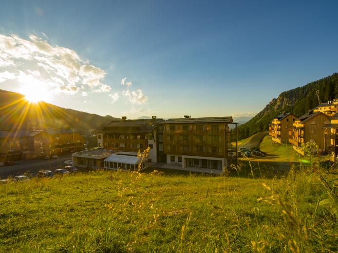 Das Almhotel Kärnten erstrahlt im warmen Licht der aufgehenden Sonne. Im Vordergrund blüht eine Wiese, während sich das moderne Hotel harmonisch in die idyllische Alpenlandschaft einfügt. Im Hintergrund ragen bewaldete Hänge in den klaren, blauen Himmel hinauf. Das Almhotel Kärnten am Nassfeld