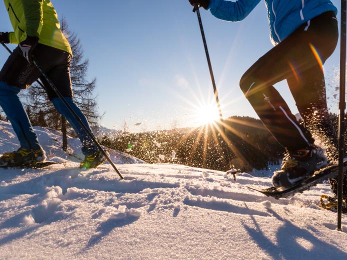 Dynamische Szene von zwei Personen beim Schneeschuhwandern durch frischen Pulverschnee im Sonnenlicht. Die tiefstehende Sonne taucht die verschneite Winterlandschaft in warmes Licht und sorgt für eine idyllische Atmosphäre. Perfekt für Winterurlaub in Kärnten. Schneeschuhwandern am Nassfeld in der Nähe des Almhotel Kärnten