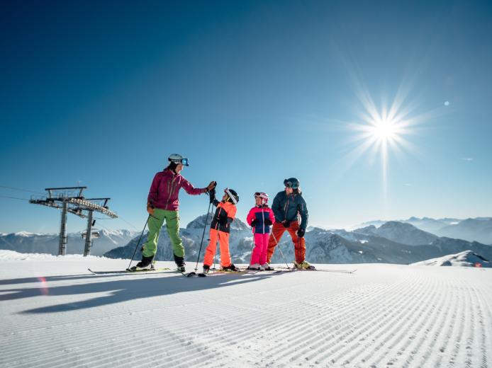 Eine Familie genießt gemeinsam einen sonnigen Skitag auf perfekt präparierten Pisten in den Kärntner Alpen. Im Hintergrund erheben sich eindrucksvolle Berge unter strahlend blauem Himmel. Die Szene vermittelt Freude, Naturverbundenheit und ideale Bedingungen für Wintersportfreunde. Familien-Skierlebnis am Nassfeld in Kärnten nahe dem Almhotel Kärnten